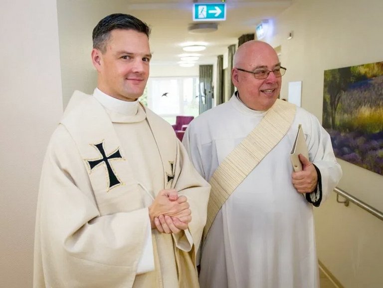 Pater Jörg und ein weiterer Pater in einem Gang einer Einrichtung lächeln erwartungsvoll.