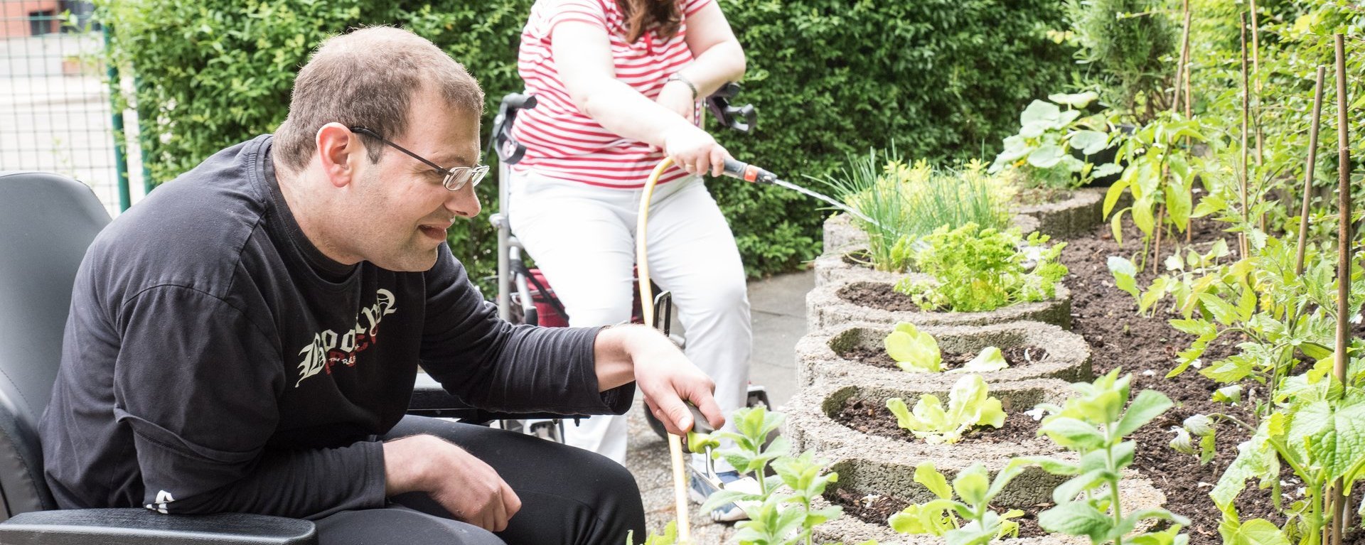 An einem Hochbeet arbeiten ein Mann und eine Frau. Der Mann hat eine kleine Gartenschaufel in der Hand, die Frau im Hintergrund gießt mit einem Gartenschlauch die Pflanzen.