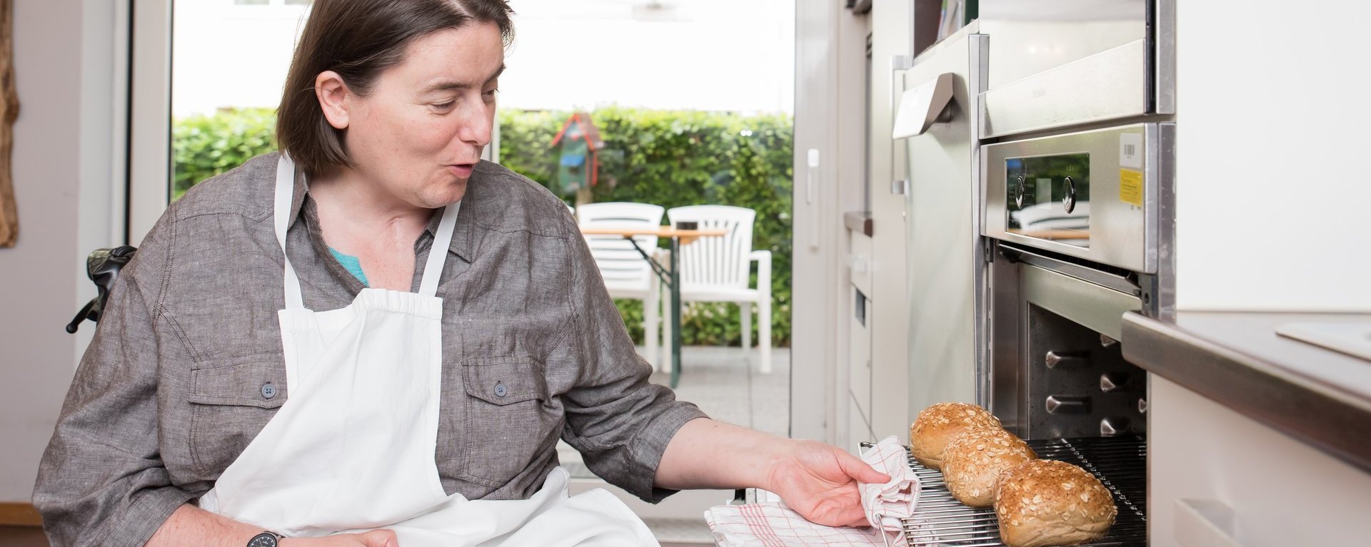 Eine Frau im Rollstuhl holt in einer Küche einen Backrost mit frischen Brötchen aus dem Ofen.