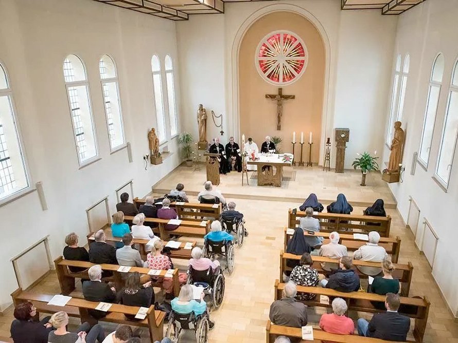 In einer Kirche blickt man von Oben auf die Besucher und den Altar neben dem vier Personen sitzen.